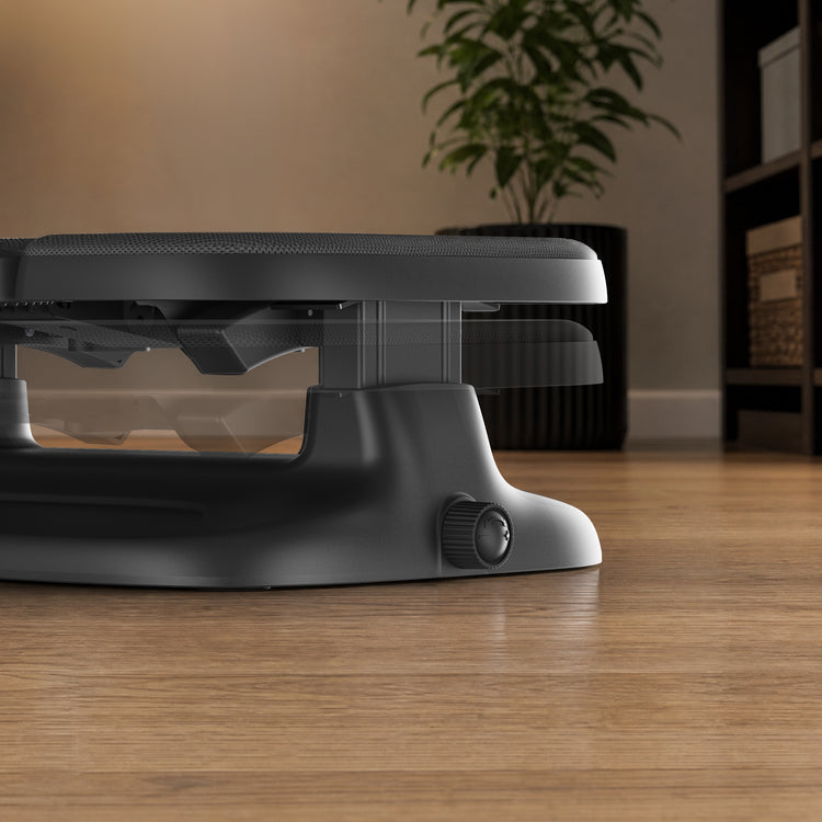 A close-up of an ergonomic foot rest on a wooden floor in a home setting, with a plant and shelving visible in the background.