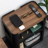Wooden side table with remote control, phone, and other items on a gray carpeted floor.